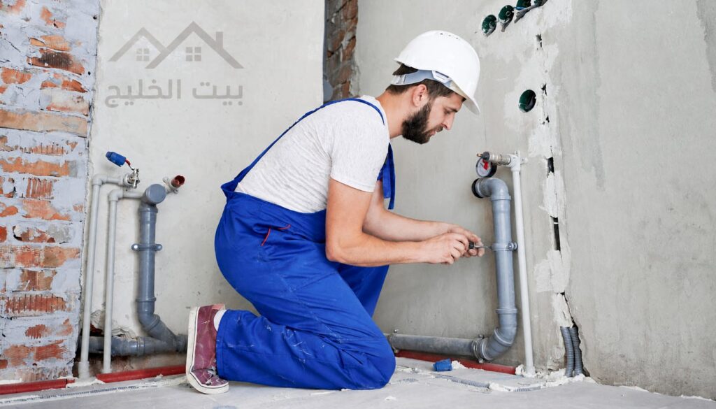 فني صحي بمشرف يقوم بتوصيل خطوط المياه داخل الجدار عبر المواسير، مع إشارة إلى خدمة الطوارئ في جمعية بيان بالكويت
